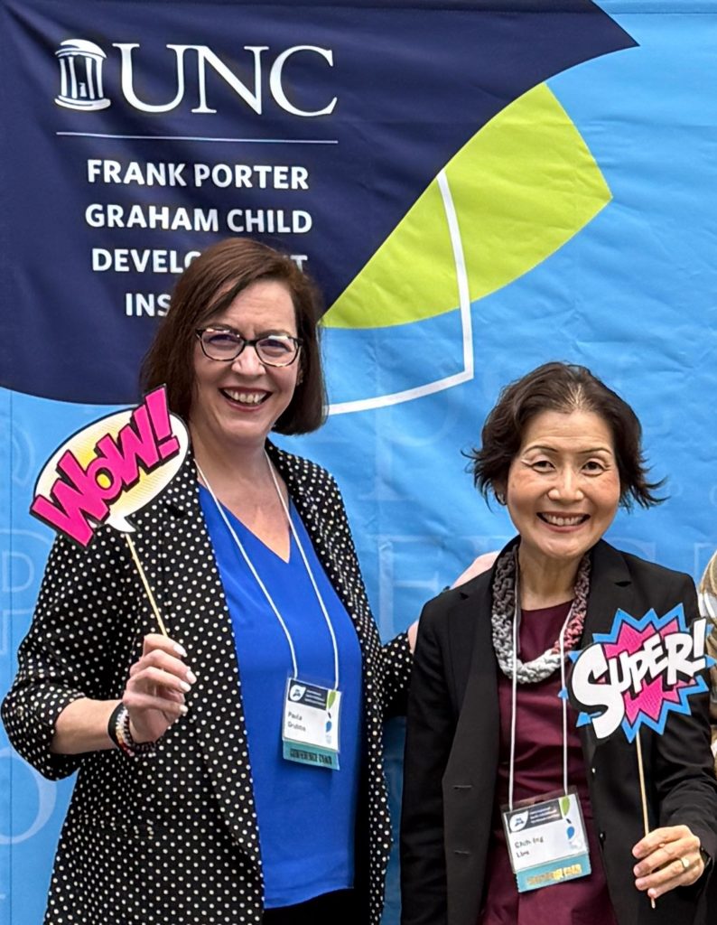 Paula and Chih-Ing smile, standing in front of a UNC Frank Porter Graham Child Development Institute banner. Paula holds a speech bubble sign that says, "Wow!" and Chih-Ing holds a sign that says, "Super!"